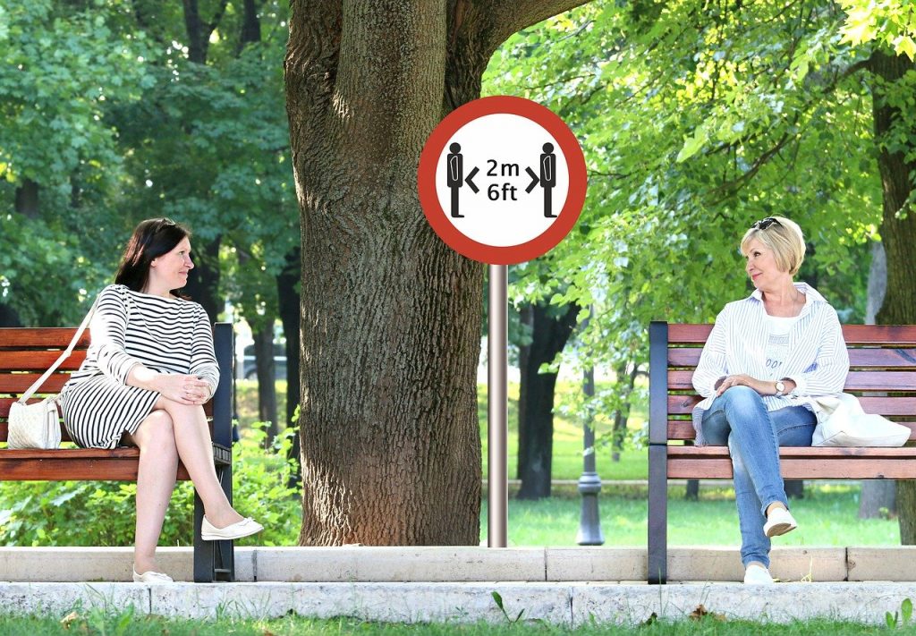 An image of two women sitting on benches in a park with a two metre apart social distancing sign between them